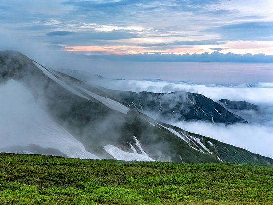 Nebel steigt zwischen grüner Anhöhe und schneebedecktem Gipfel empor