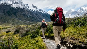 Zwei Wanderer auf einem schmalen Pfad durch grün bewachsene Landschaft mit schneebedeckten Bergen im Hintergrund