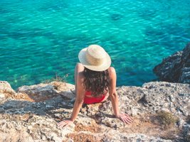 Frau mit Hut sitzt auf einem Felsen und schaut ins türkisblaue Meer