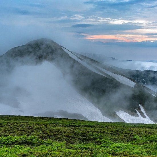 Nebel steigt zwischen grüner Anhöhe und schneebedecktem Gipfel empor