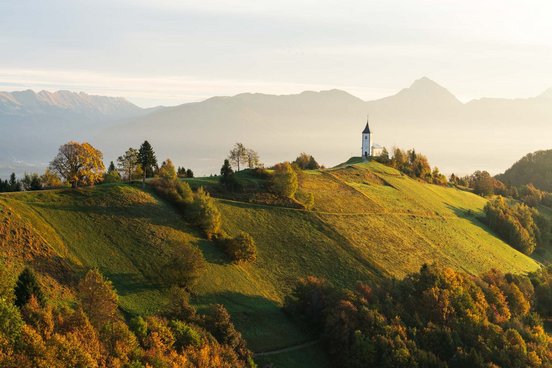 Kirche auf grünem Hügel im Morgenlicht mit Bergen im Hintergrund