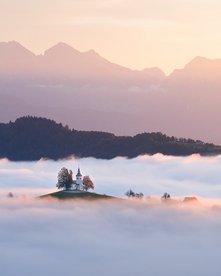 Kirche auf einem Hügel ragt aus Nebelmeer mit Gebirge im Hintergrund beim Sonnenaufgang